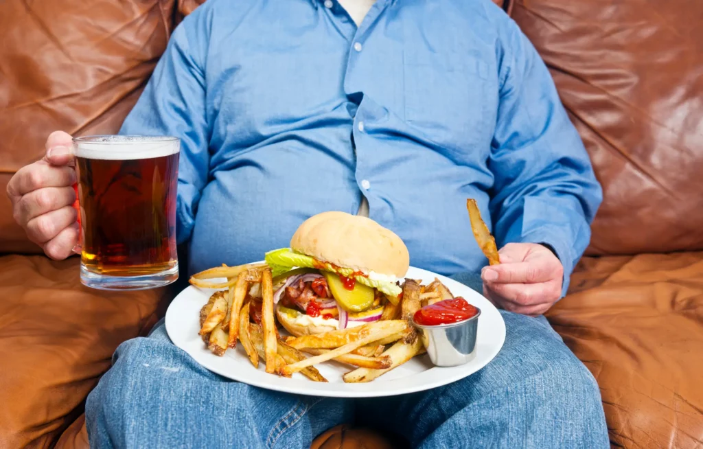 A photo of an overweight man sitting on an old couch with a very large unhealthy meal on his lap and a pint of beer in his hand. Obesity is a major cause of diabetes.